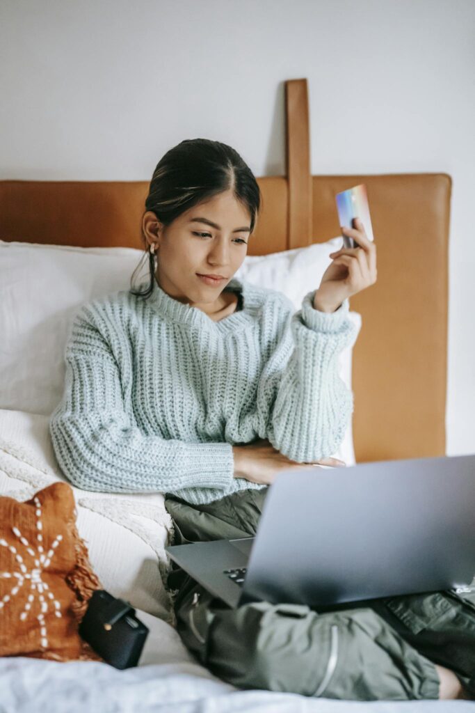 A young woman relaxes in bed, shopping online with a laptop and credit card in hand.